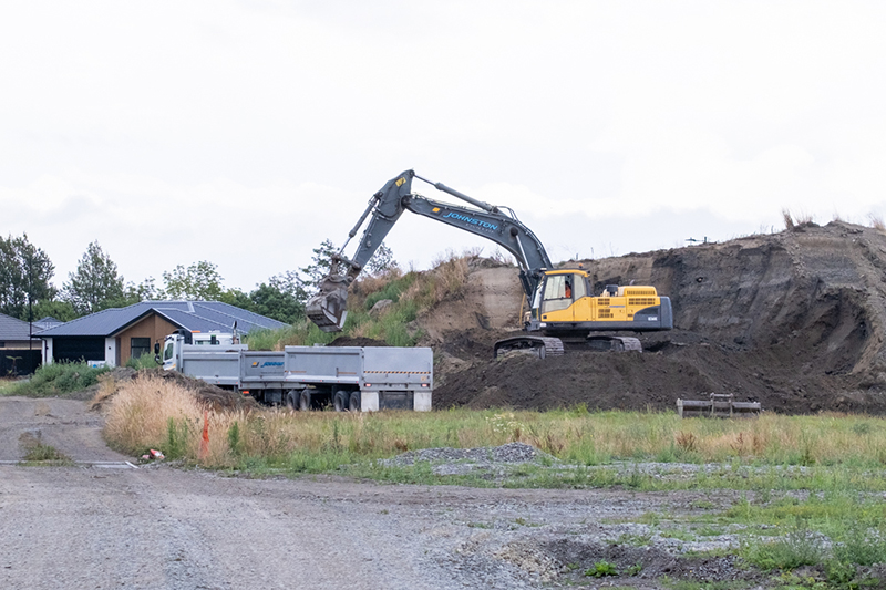 A yellow digger is moving soil from a large pile to the back of a truck. There is a house in the background.