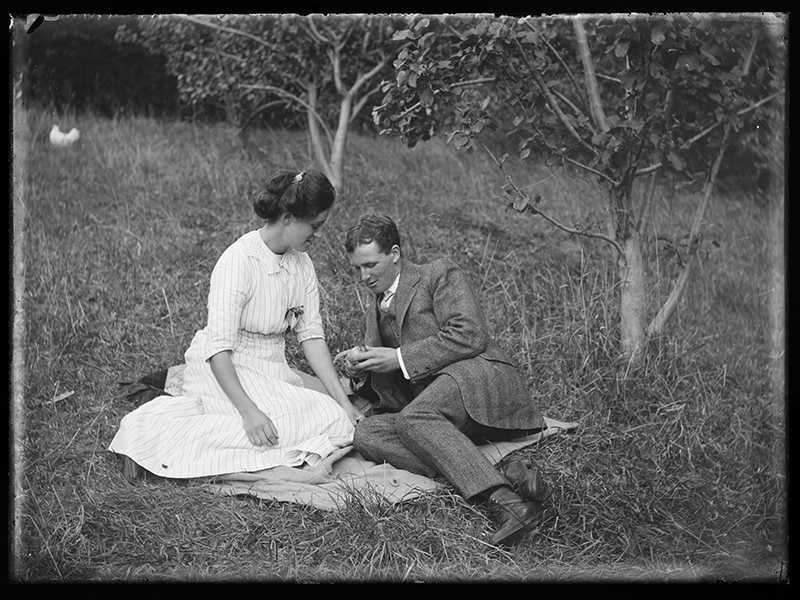 A black and white photograph of a woman and man sitting on a picnic blanket in a grassy field.