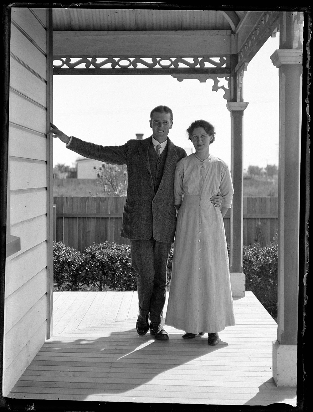 A black and white photo of a man and a woman standing close together and looking at the camera on a verandah.