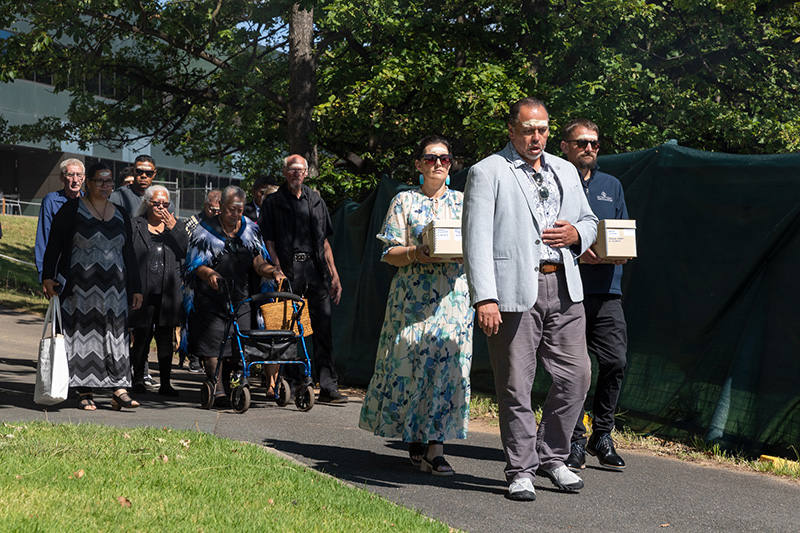 Several people walking along a path. Two are carrying boxes and walking behind the one person in front.