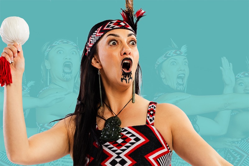 A woam with a chin tattoo and cultural dress is holding a poi (ball on a string) in the air with one hand. She is mid chant and is performing before an unseen audience. There is an opaque photo in the background of three other women doing the same thing.