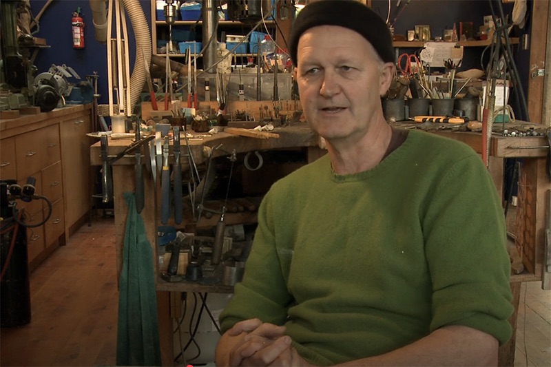 A man in a woollen cap and green jersey is talking to someone just off camera. In the background is his jewellery studio.