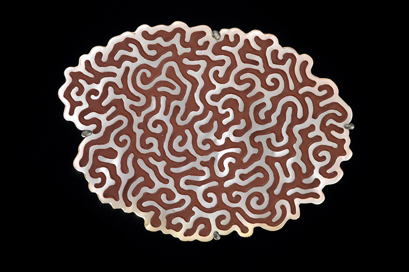 A brooch on a black background. It is made from a piece of shell with rounded maze-like shapes carved into it which have been filled with red lacquer, making it look like a stylised brain.