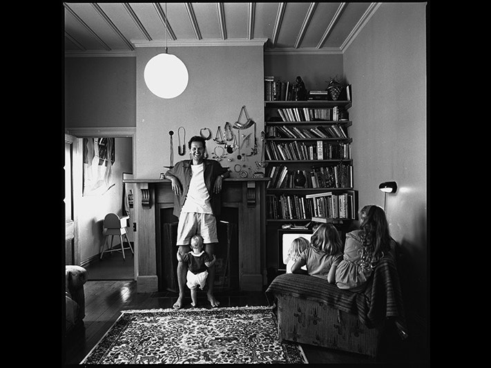 Warwick Freeman frame 10, by John Daley, about 1987. Gift of John Daley. Te Papa (F.012323/10) A black and white photo of a man facing the camera with his elbows resting on the mantle of a fireplace. He has a toddler holding onto both legs, and there are two people on a chair with their backs to the camera.