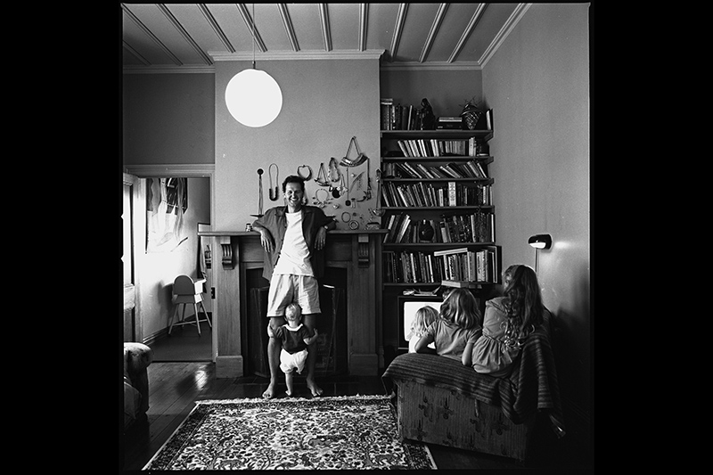 A black and white photo of a man facing the camera with his elbows resting on the mantle of a fireplace. He has a toddler holding onto both legs, and there are two people on a chair with their backs to the camera.