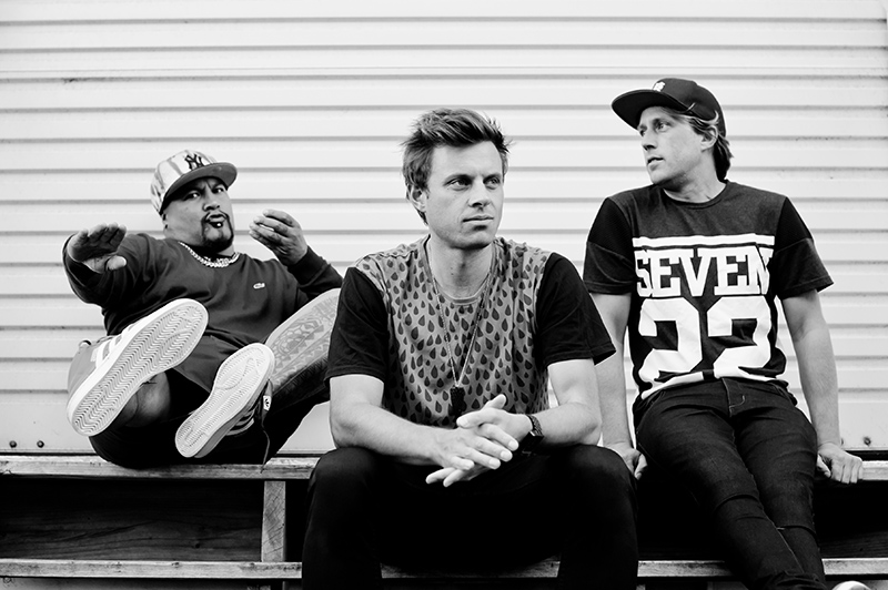 A black and white photo of three men sitting on a bench sharing chill vibes. Two are looking in different directions, and one is pulling a face at the camera with his feet in the air.