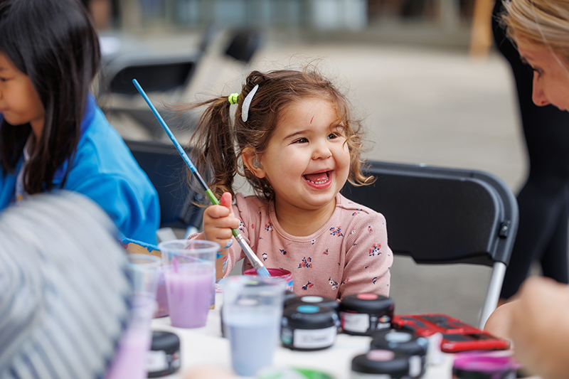 A close-up image of little girl, aged around three years old, holding a paint brush while smiling broadly at a lady to her right.