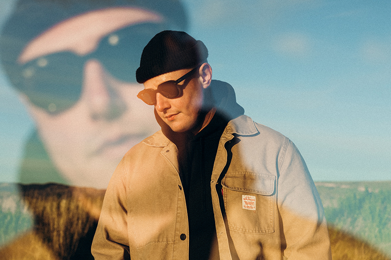 Musician Louis Baker stands in a jacket and sunglasses, with green field as his backdrop, exuding effortless style. The photo has a close up of his face in sunglasses and a beanie super-imposed over it.