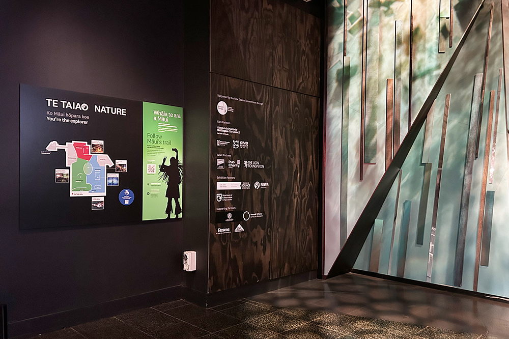 A photo of two signs at the beginning of a museum exhibit. One of the signs has a map of the space and a QR code to download an app. And the other sign has all the sponsor logos.