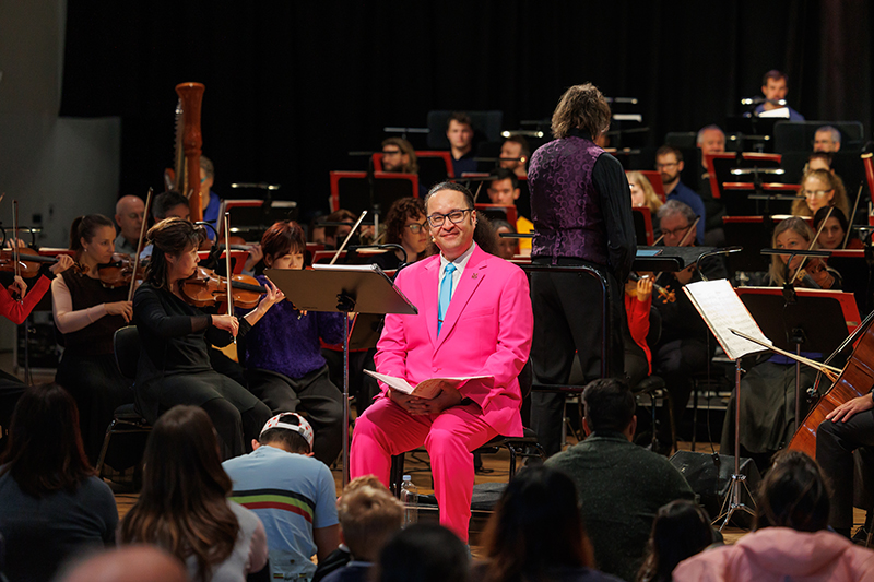 A man with his hair tied back and wearing a bright pink suit is sitting in front of an orchestra. There are kids sitting on the floor looking at them.