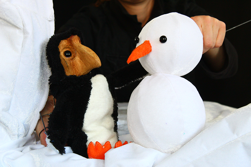 A penguin puppet and a snowman puppet are sitting on white cloth which is imitating snow.