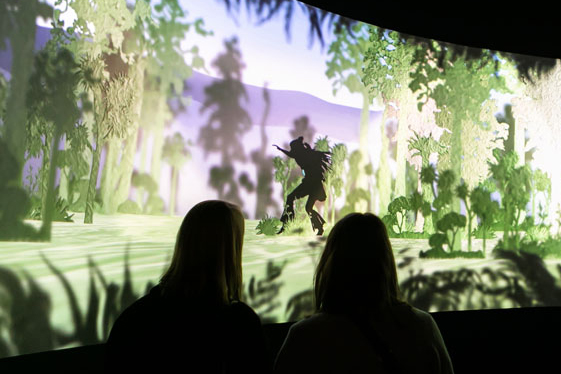 Two silhouetted people are watching a screen where another silhouetted person is creeping through the forest.