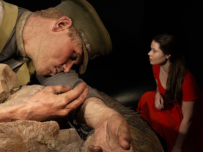 Visitor with a large scale model in Gallipoli: the scale of our war exhibition, 2015. Photo by Michael Hall. Te Papa (63315) A woman in a red dress is crouched next to a giant model of a soldier.