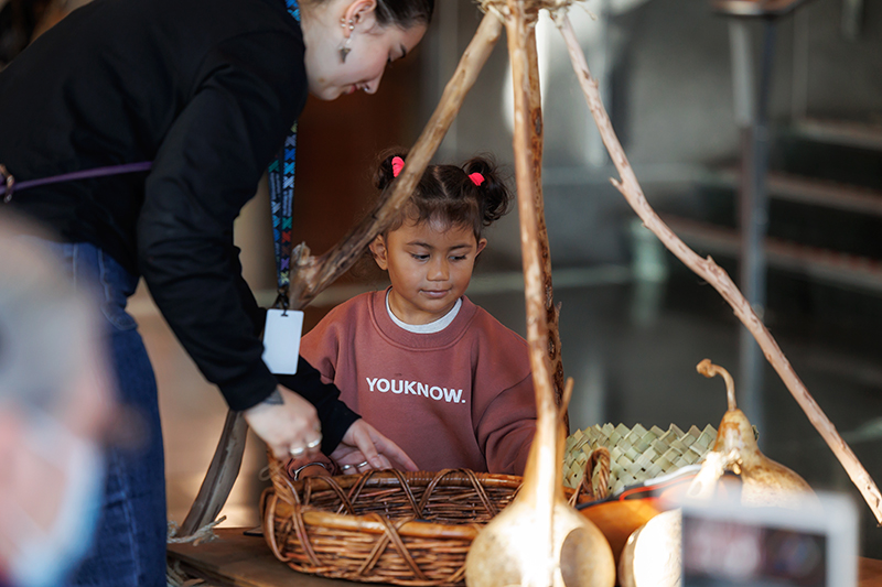 A woman is bending down to reach a basket that is sitting under a wooden structuire. A small child is standing beside her and looking at the basket.