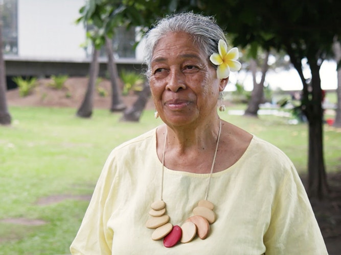 Still from video of Sulieti Fieme'a Burrows interview. Te Papa An older woman is standing on grass smiling at someone off camera. She is in a yellow top and is wearing a frangipani flower behind her left ear.