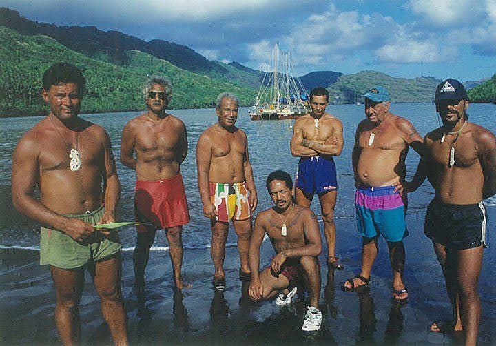Te Aurere crew members with Nuku Hiva locals, Marquesas, 1995. Courtesy of Te Tai Tokerau Tarai Waka Inc and Jack Thatcher Seven men wearing only shorts are standing on the water's edge. There is a double-hulled vessel in the harbour behind them.