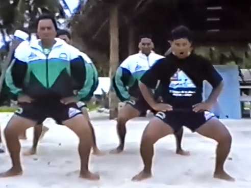 A still from Haka: ‘E Kau ki te Tai’ performed by the crew of Te Aurere on their voyage to Rarotonga. Several men are mid-haka or ceremonial dance on a sandy beach. The image is blurry as it is from old footage.