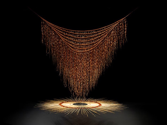 Maureen Lander, Huna: Kaitiaki o te Harakeke, 2012. Installation view, 2025. Commissioned by Te Papa (ME024116) A black rectangle with an artwork of a woven cloak hanging over a circle of similar woven material.