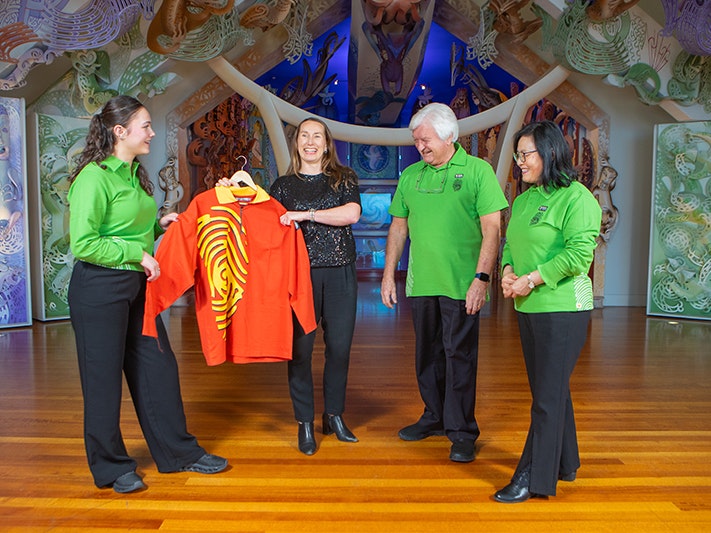 Te Papa Tumu Whakarae | Chief Executive Courtney Johnston with a “fun shirt” from the opening of Te Papa, with Hosts wearing the new Host uniform launched today. Photo credit: Te Papa. Three people in green shirts and one person is in a black shirt. Two of them are holding another shirt that is yellow and orange.