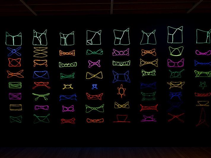 Maureen Lander, String games,1997. Installation view (detail), 2025. Commissioned by Te Papa (1999-0033-1) A black wall with eight columns of neon-coloured patterns created with string.