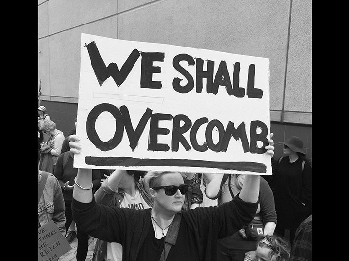 We Shall Overcomb placard held by Johanna Mechen at the Women's March in Civic Square, Wellington, 21 January 2017, New Zealand, by Adam Dransfield. Gift of Johanna Mechen, 2017. CC BY-NC-ND 4.0. Te Papa (O.051294) A black and white photo of a woman holding a large placard that says "We shall overcomb".