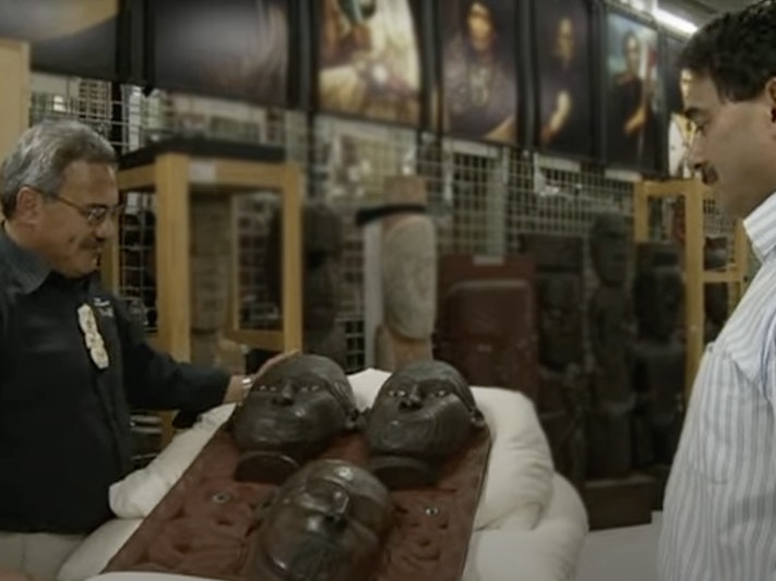 Still from Tales from Te Papa. Tā Moko Panel Two men are standing either side of a carving that is laid on a pillow. There is a museum store space in the background.