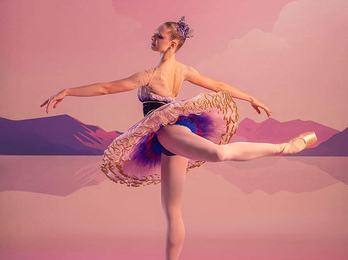 Soloist dancer Jemima Scott, July 2025. Photo by Ross Brown. RNZB A ballerina in arabesque position, wearing a pink, purple and gold sugar plum fairy costume with a tiara. The backdrop is a pink sky and purple mountains.
