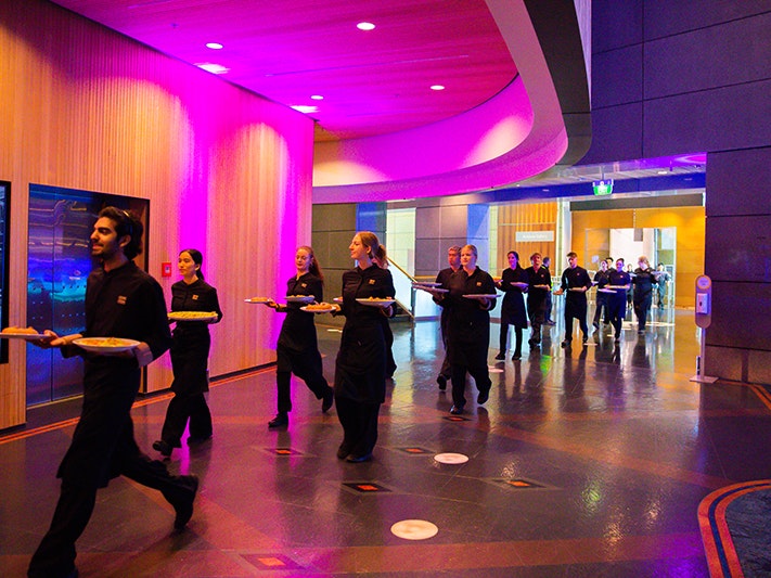 Rebel Hearts Galentine's Dinner, 2025. Photo by Fineline Photography. Te Papa (257771) Several people dressed in black are carrying platters of food through an open foyer in a large building.