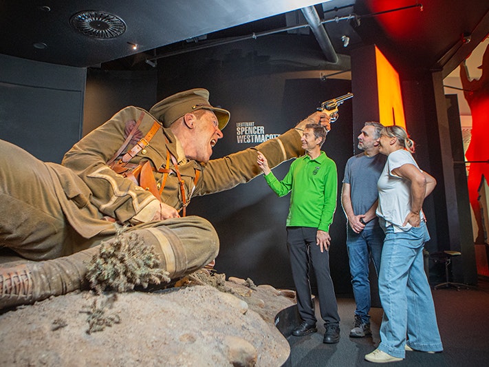 Te Papa 2025 Tour Stock. Photo by Jeff McEwen. Te Papa (270465) A man in a green shirt is pointing to a giant model of a soldier while talking to two other people.
