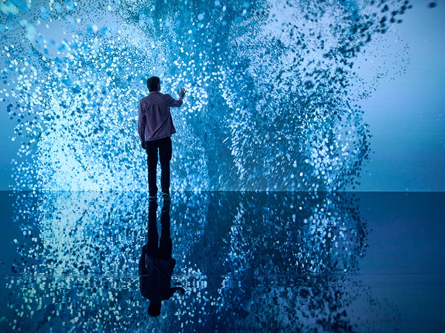Marshmallow Laser Feast: Works of Nature, 2023, ACMI. Photo © Eugene Hyland A person is standing in front of a giant screen that has abstract light patterns on it. They have their hand out to it and it is all reflected on a shiny floor.