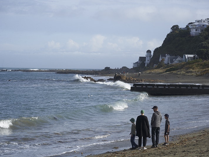 Island Bay. Photo by Jean-Claude Stahl. Te Papa Two adults and two children are walking along a beach. There are some houses perched on a hill in the background.