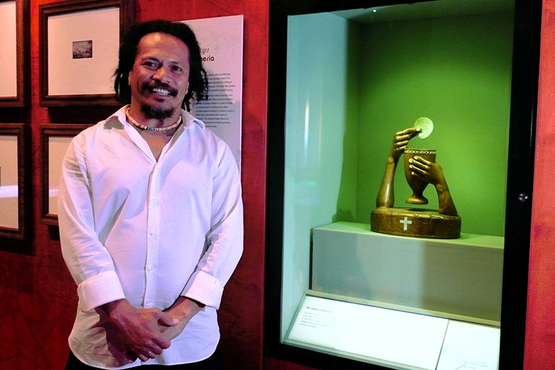 Selwyn Teho and 'The Great Elevation' chalice and host. Photo by Rowan Moulder. Te Papa A dark skinned Solomon Island man in a white dress shirt stands beside a carved sculpture. The sculpture is dark timber chalice and 2 arms, inlaid with shell. One arm holds the chalice and the other holds a communion wafer made of shell.