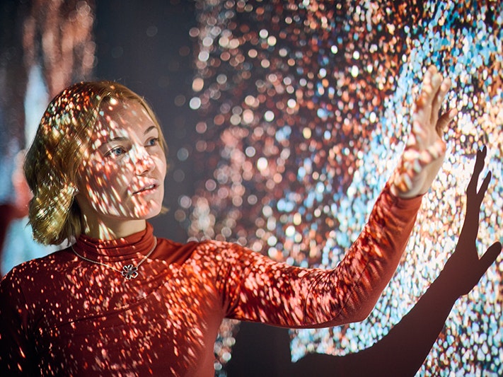 Marshmallow Laser Feast: Works of Nature, 2023, ACMI. Photo © Eugene Hyland. A woman has a lightshow playing over her face and she has an arm up near the screen it is projecting on.