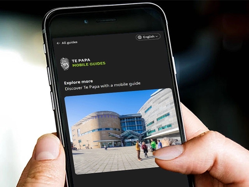 Te Papa mobile guide in English. Te Papa A close up of a hand holding a mobile phone that has an image of Te Papa from the front forecourt.