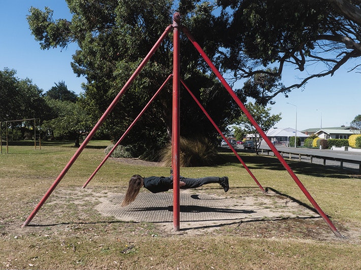 Nela Fletcher, Fitting in #189 Otakaro Park, 2022. Purchased 2025. Te Papa (O.052237) A person with long hair is lying flat on a swing in a park. It looks like they are hovering above the ground.