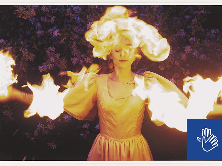 Tia Ranginui, The tangi of Rūaumoko, 2023. Purchased 2024. Te Papa (O.052215) A woman wearing a silk gown with puffy sleeves stands with arms outstretched in front of a background of purple flowers. Flames engulf her head and arms while she smiles serenely with eyes closed.