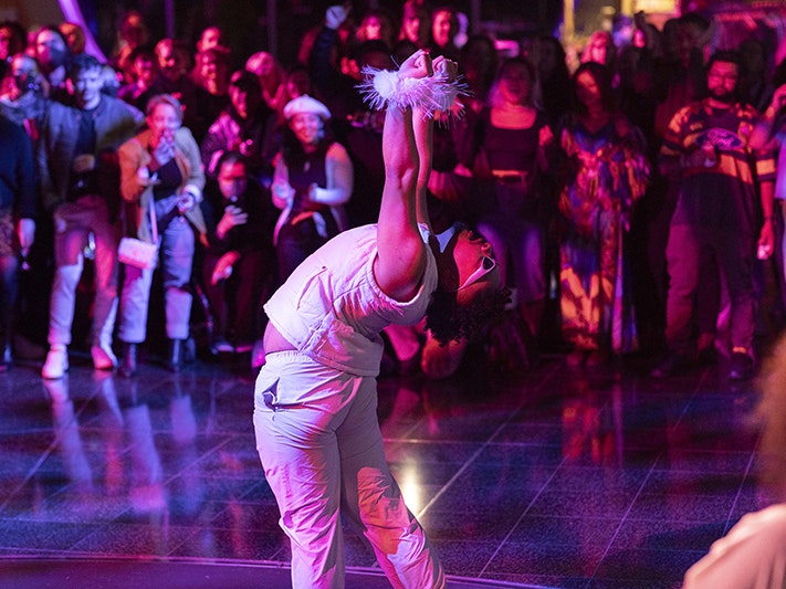 Matariki After Dark 2024. Photo by Neil Price. Te Papa (245644) A person in a white top and trousers, with white fur around their wrists is performing in front of a crowd under red and blue lights.