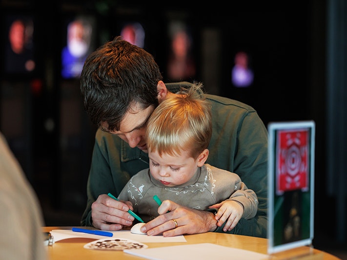 Matariki Whānau Day 2025. Photo by Jo Moore. Te Papa (268881) An adult has a toddler on their knee and they are both colouring in a piece of paper on the table in front of them.