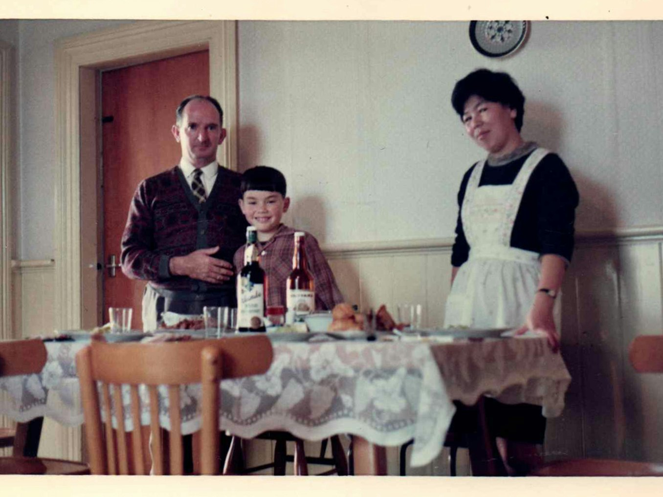 Setsuko, Mick, and Pat preparing for a meal. Photo courtesy of Jo Cullen A father, mother and child are posing behind a dining table.