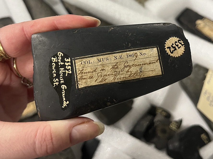A toki, or adze, from the Collection store. Photo by Manon Verdello. Toki (adze blade), carver unknown, Wellington. Gift of Mr Huntley, 1869. Te Papa (ME003352) A hand is holding a stone adze with old museum stickers and handwritten labels on it.