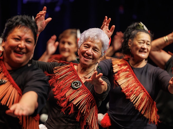 Tūhourangi Ngāti Wāhiao Koeke, Taikura Kapa Haka 2025. Photo by Jo Moore. Te Papa (270891) Three women in front of other women performing a song with hand actions. They all have the same red sash on.