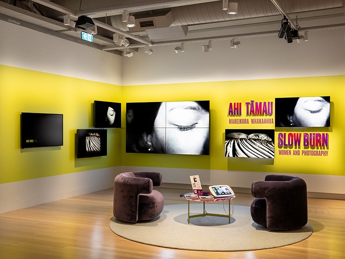 Entrance to Ahi Tāmau | Slow Burn, 2026. Photo by Maarten Holl. Te Papa (278129) 2 low, cushioned, crushed velvet armchairs with rounded half backs next a small touch screen on a low round table atop a round rug on hardwood floor. 5 bright screens are mounted on the back corner walls. The exhibition name Ahi Tāmau | Slow Burn is in uppercase purple and red letters coloured against a chartreuse rectangle. The screens and seating are under spotlights causing strong shadows.