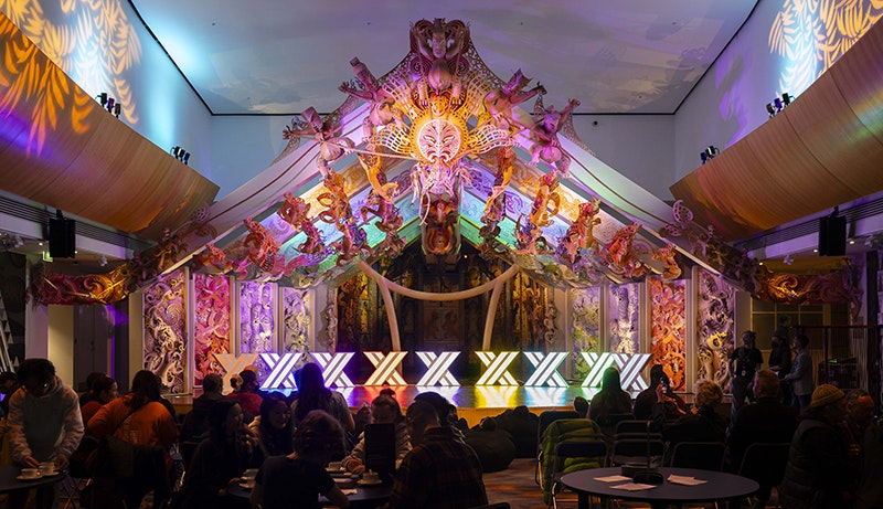 Matariki After Dark 2024. Photo by Neil Price. Te Papa (245519) A room with coloured lighting and colourful carving behind the stage. On the stage are nine lights in x shapes.