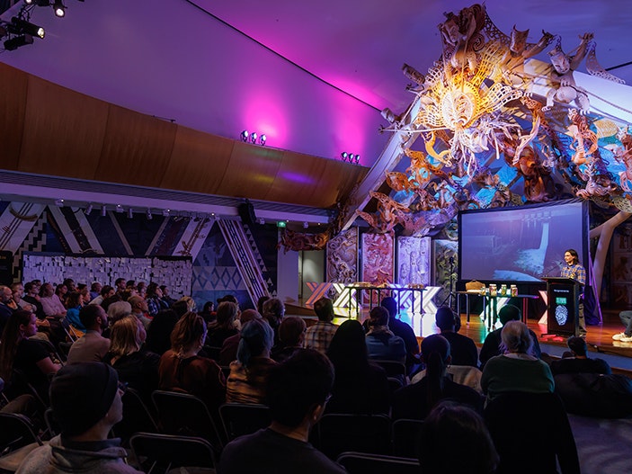 Matariki Kōrero: Waitī 2024. Photo by Jo Moore. Te Papa (244995) A person is standing on a colourful stage with a display screen behind him. He is talking to an audience.