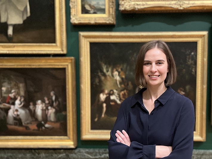 Dr Victoria Munn. Photo supplied by speaker A woman is standing with her arms crossed in front of paintings hung in a gallery.