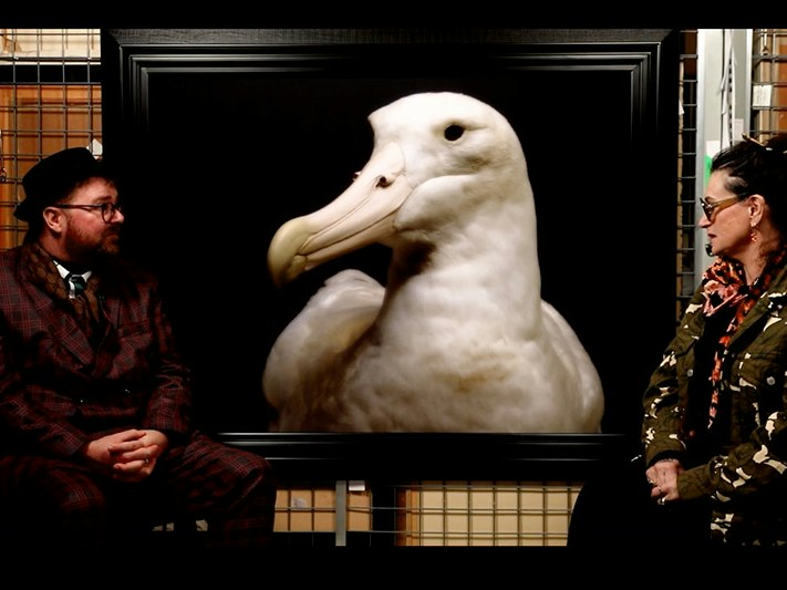 Still from Andrew Paul Wood talking to Fiona Pardington. Aigantighe Art Gallery Two people are sitting facing the camera, but talking to each other in front of a large photo of an albatross.