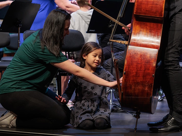NZSO at Te Papa October 2023. Photo by Jo Moore. Te Papa (236794) A woman is kneeling on the ground showing a child a cello.