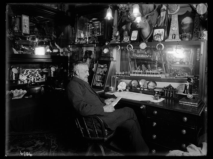 Captain Hennah, Inspector of Messengers, Government Buildings, 1913, Wellington, by James Ingram McDonald. Te Papa (MU000523/001/0426) A black and white historical photo of a man in a very crowded office reading something in his hands.