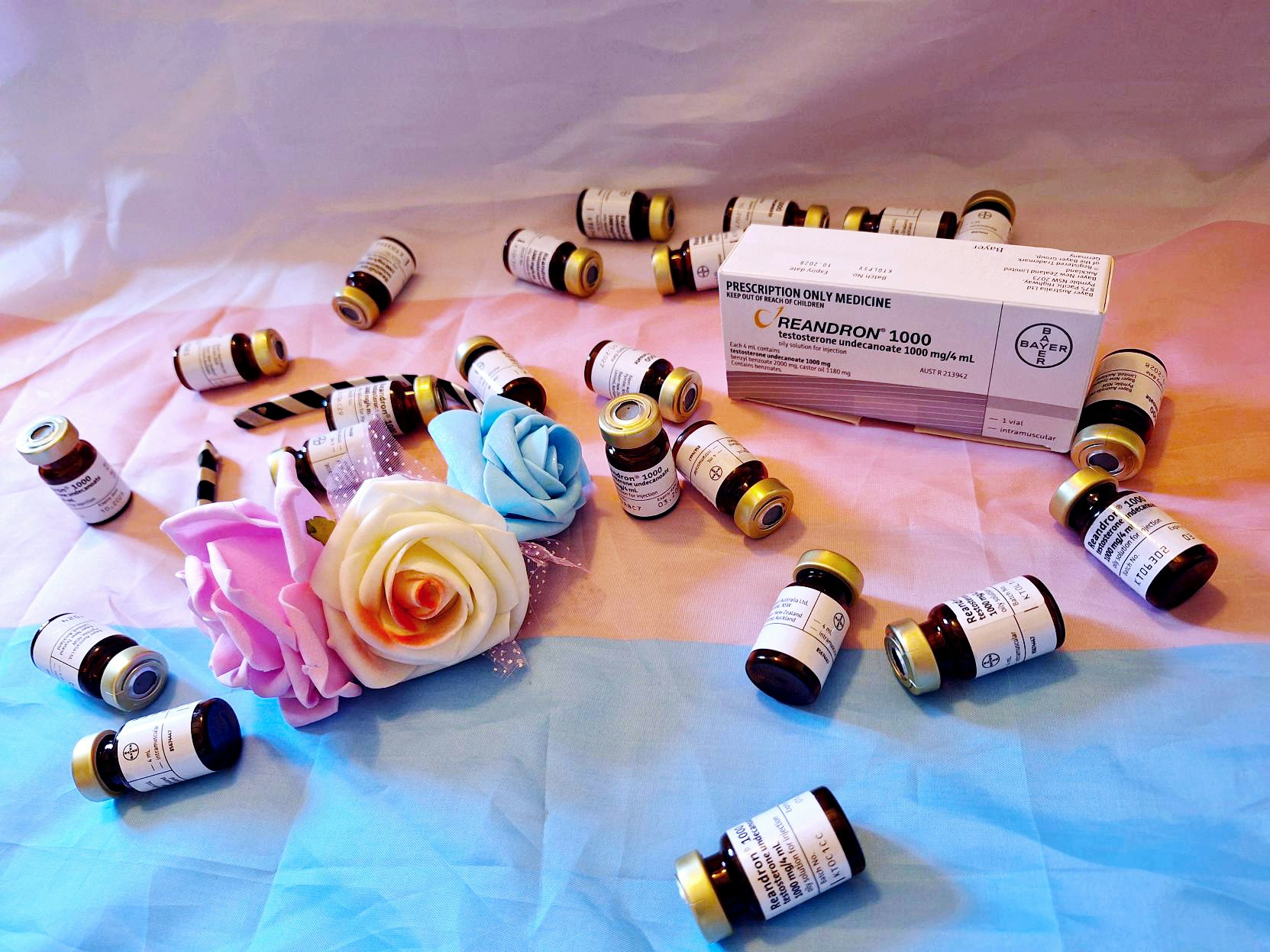 "What it takes to be a man - 8 years of HRT". Photo by Rowan Moulder, 2026. Te Papa. 22 small brown, gold capped vials with white labels are scattered across a white, pink and blue striped transgender flag. A headband with foam pink, white and blue roses lies next to the white, rectangular box of Reandron, a common testosterone hormone replacement therapy.