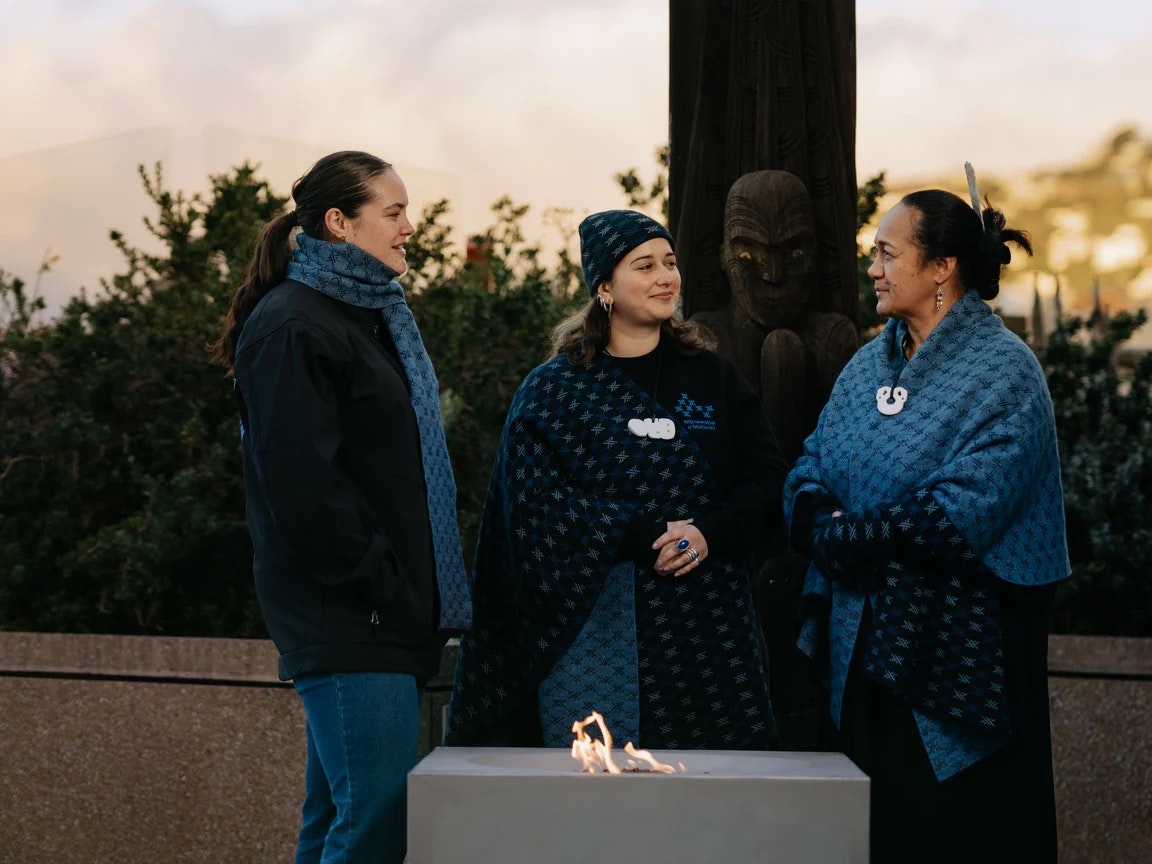 Te Papa Matariki Merchandise 2026. Te Papa Three women in clothing with the same design are standing next to a fire on a balcony.